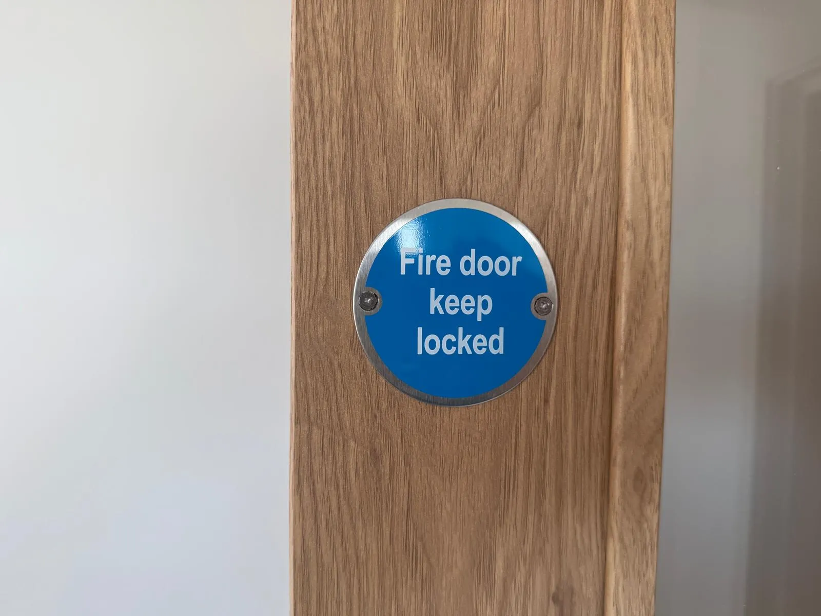 A round blue Fire Door Keep Locked sign fixed to an oak-veneered timber door, one of the visible markings used to help identify a fire door